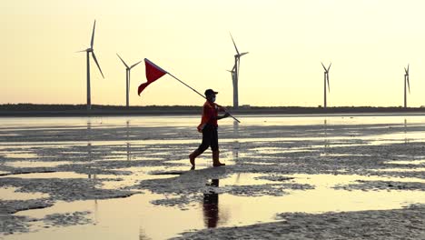 un guardacostas sosteniendo una bandera roja, caminando por las llanuras de marea con turbinas eólicas girando y girando a lo largo del horizonte durante la hermosa puesta de sol en horas doradas, humedal gaomei, taichung, taiwán