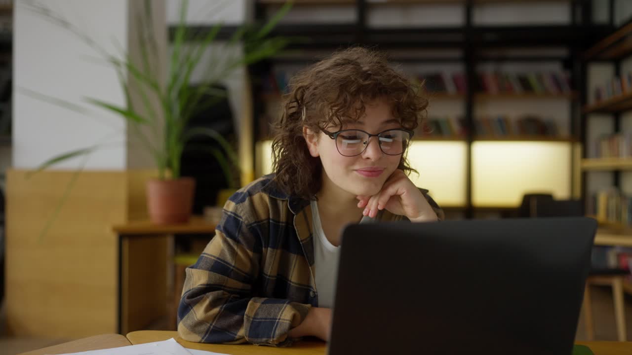 una chica feliz con cabello rizado y gafas se comunica a través de video con sus compañeros de clase usando una computadora portátil en la mesa de la biblioteca