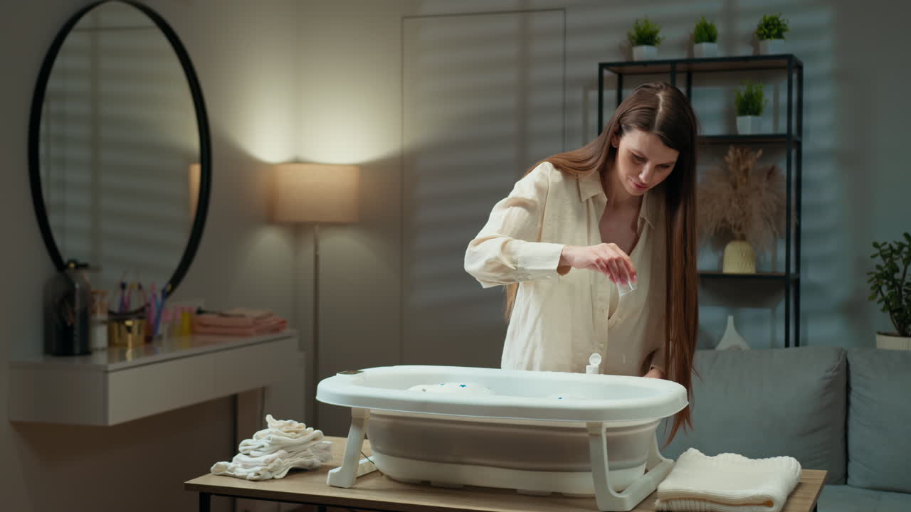 Woman carefully adds liquid soap to portable baby bathtub filled with water and bubbles mother