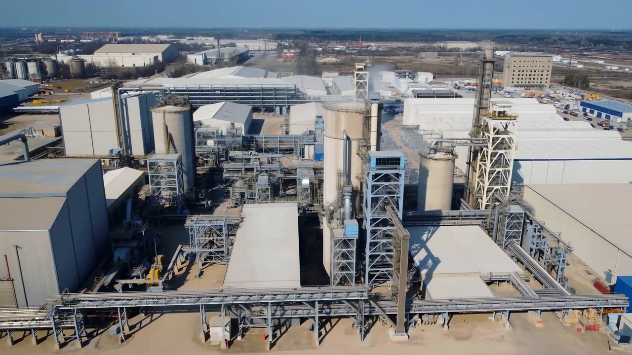 High oblique aerial view shows expansive industrial processing plant with silos, chimneys, lattice towers, conveyors and flowing vent plume above central production core in clear daylight