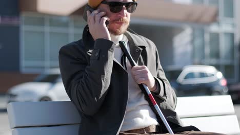 un hombre con discapacidad visual hablando por teléfono, sentado en un banco en la ciudad.