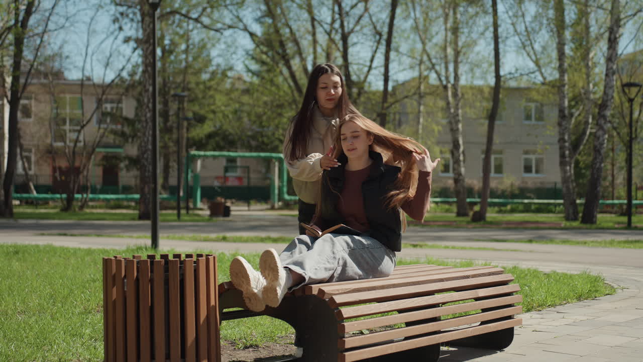 Escena de estilismo en banco, estilismo asiático, pelo caucásico, tacto suave y ajuste de mechones en un banco soleado del parque, chaquetas informales y ambiente tranquilo de primavera, aseo natural y cuidado amable