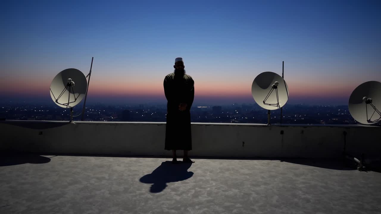 Imam stands on a rooftop at dawn, overlooking a city skyline with satellite dishes in the foreground, creating a serene and contemplative scene