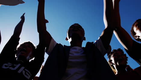 a low-angle shot of a group of young people with their hands raised against a bright blue sky, possibly at a protest or rally