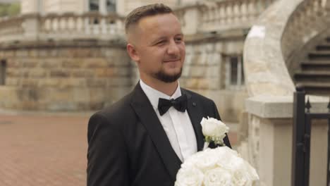 Groom-goes-down-the-alley-near-old-castle-with-a-wedding-bouquet-to-his-beloved-bride,-first-meeting