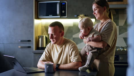 man sits at the table looking at his laptop. woman holding a cute blond baby stands near the man. kitchen backdrop.