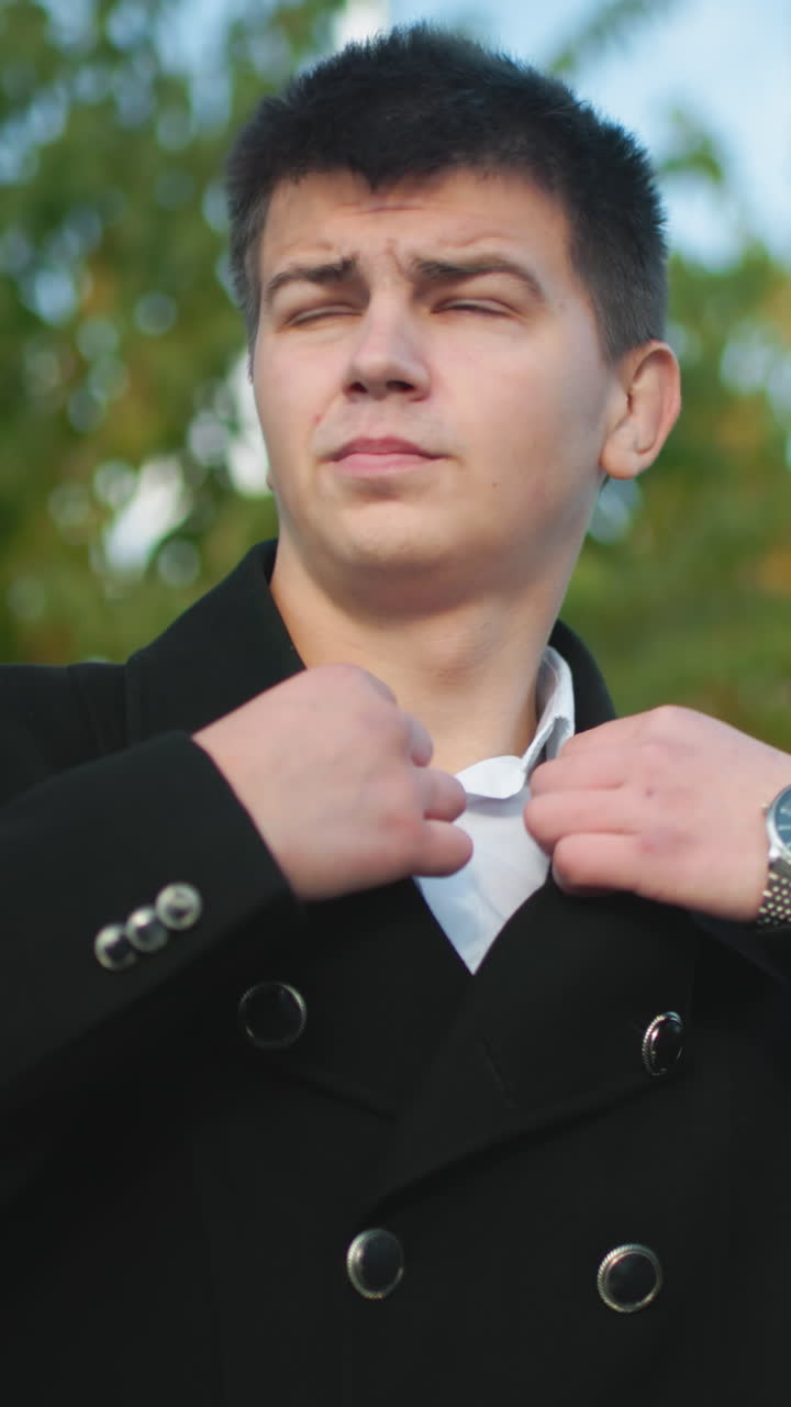 두 손으로 목걸이를 조정하는 투자자, 밖에서 전문적인 검은 코트를 입고, 은 파란 하늘 아래, 푸른 잎자루와 도시 배경의 차량으로 자신감을 가지고 비즈니스를 준비합니다.