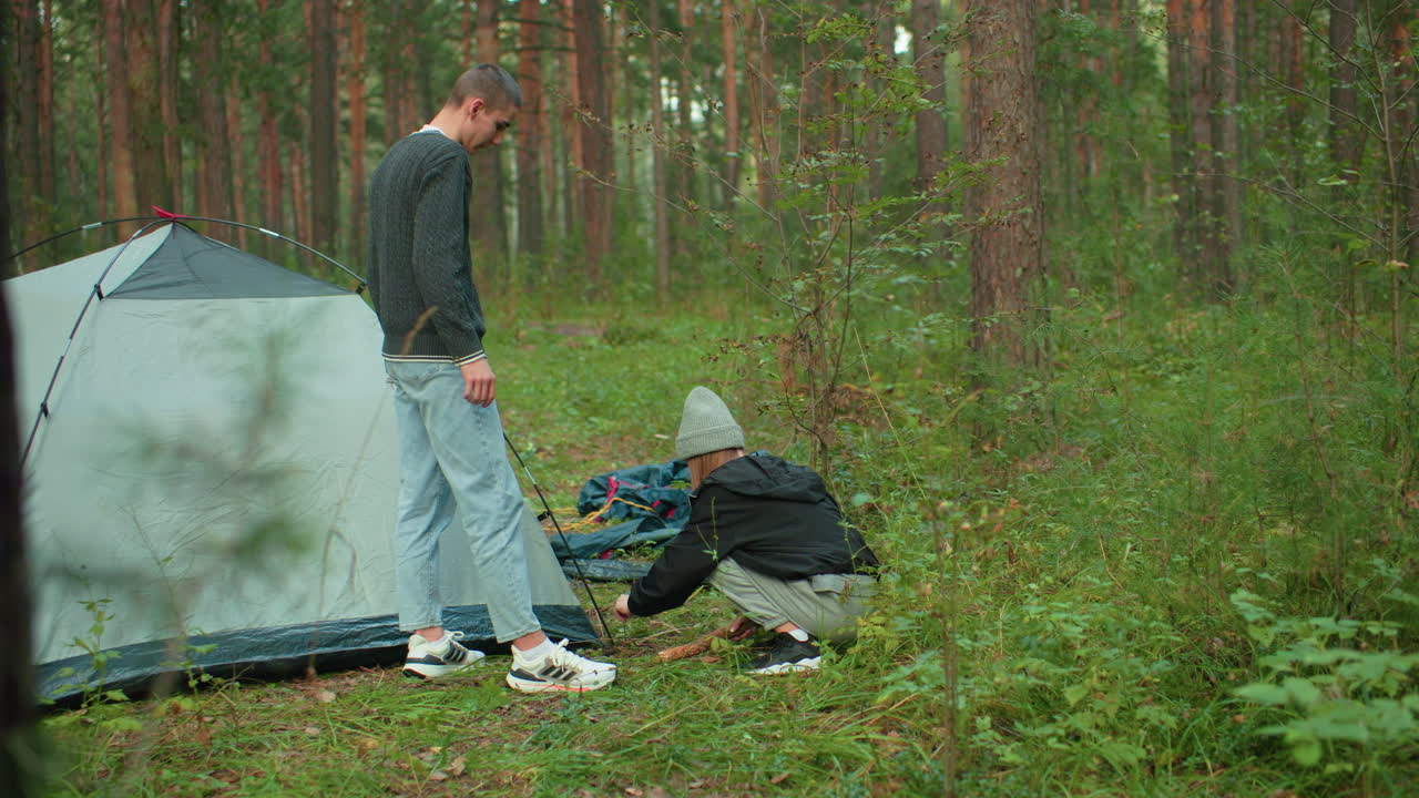 Man hands tent peg to woman squatting near tent in forest as she prepares to hammer it into ground while setting up camping shelter surrounded by lush greenery and pine trees during outdoor adventure