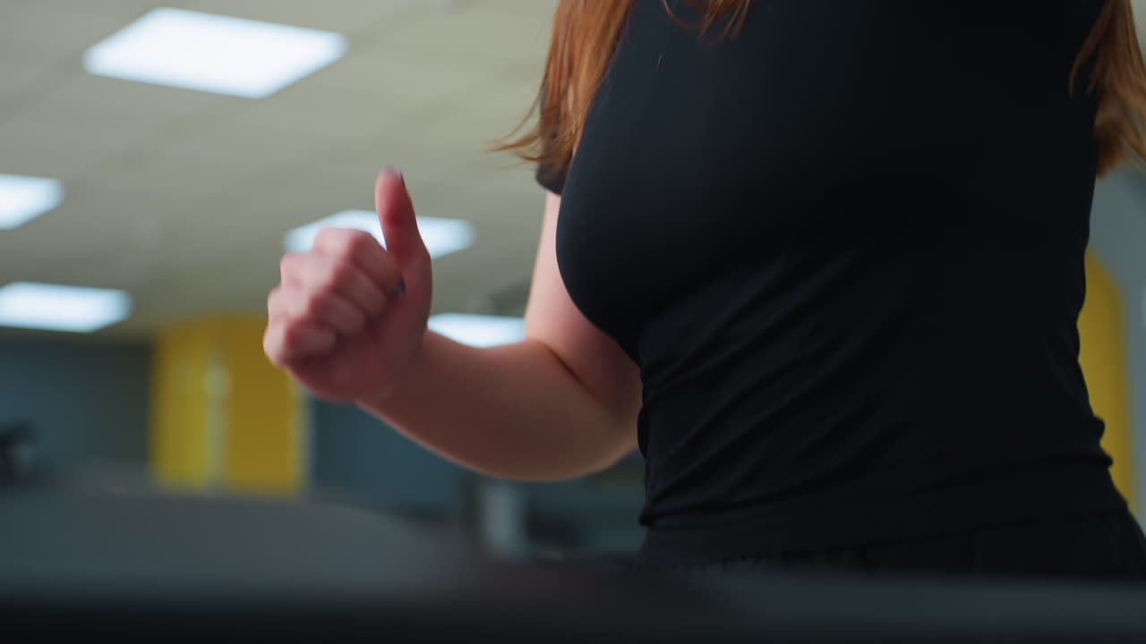 focused woman in black activewear jogging on treadmill adjusts hair mid-stride under bright indoor ceiling lights with determined expression showing workout effort in modern gym environment