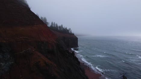 aerial view of the beautiful red cliffside that meets the atlantic ocean on the coast of cape breton island in nova scotia during a foggy and moody cold morning shot in 4k