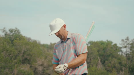 male golfer stands on the fairway holding two irons, putting on his glove as he decides which club to hit. a thoughtful, relatable moment showing game strategy and pacing during play