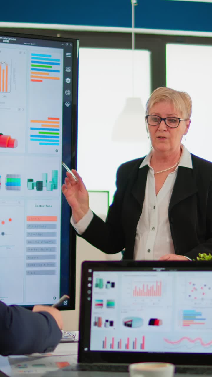 Vertical video Elderly project manager pointing at desktop presenting statistical data,