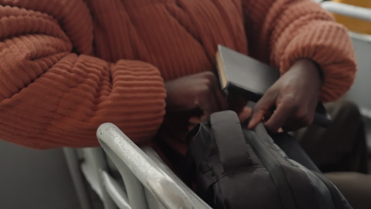 Pasajero afroamericano en autobús guardando un cuaderno, primer plano de las manos con suéter naranja en la cremallera y la tapa dura, escena tensa y concentrada, gesto protector asegurando un diario personal, roles de escritor, estudiante, autónomo