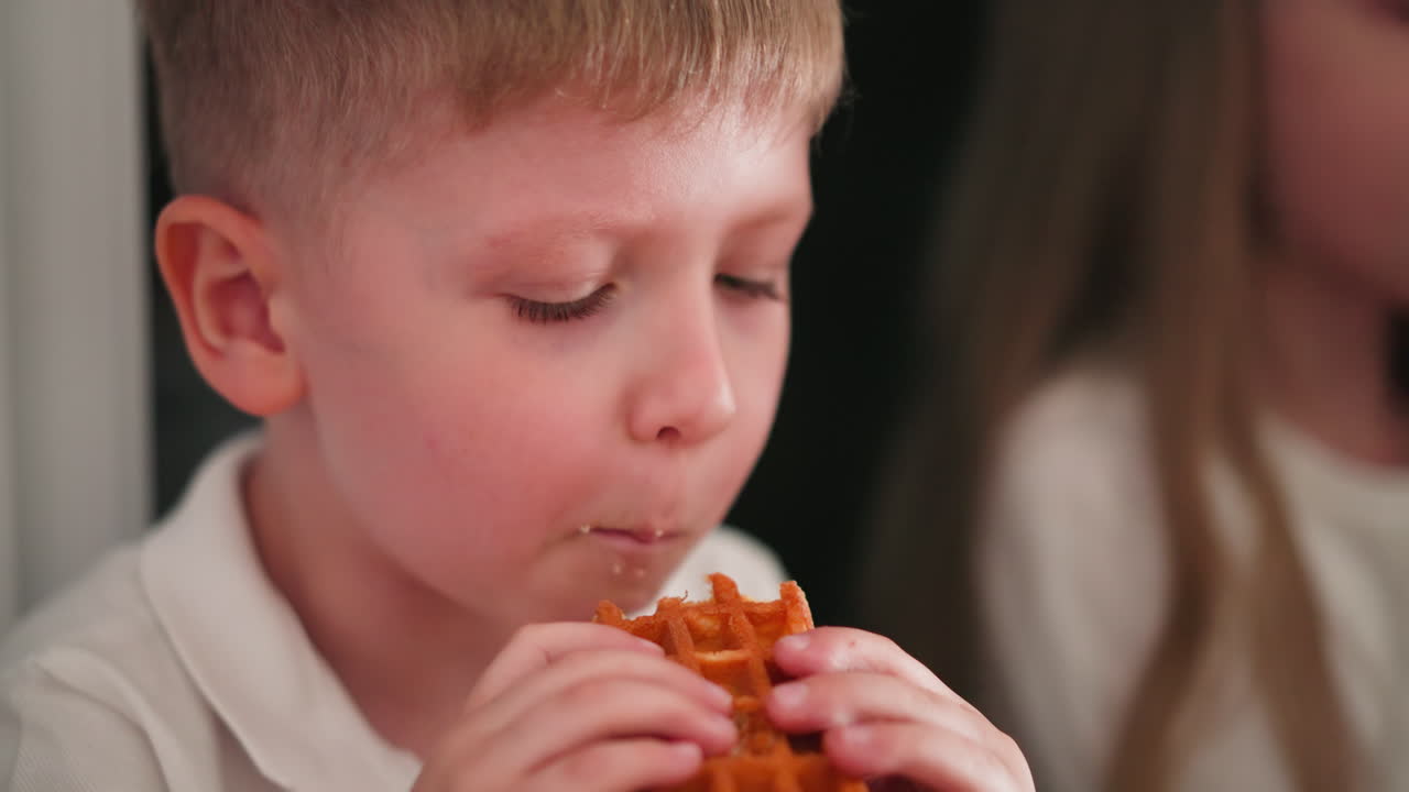 Close-up of young boy enjoying golden waffle dessert with his cousin visible in background also eating her dessert, captured in indoor setting with focused expressions and soft light
