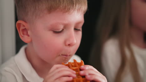 close-up of young boy enjoying golden waffle dessert with his cousin visible in background also eating her dessert, captured in indoor setting with focused expressions and soft light