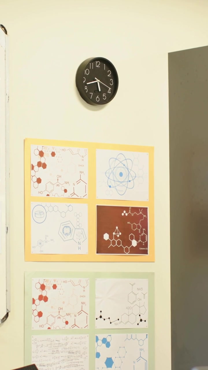 Vertical video: Panning camera shifting downward to reveal lab wall with black clock and posters
