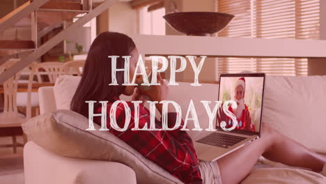 animation of happy holidays text over woman on laptop making christmas video call with santa claus