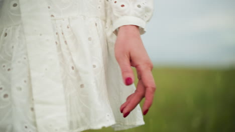 a close-up of a woman's hand with red nail polish, elegantly moving through a blurred grassy field. she is wearing a white, patterned dress, creating a soft and serene atmosphere in a natural setting