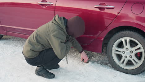 man squatting in winter jacket uses hand to lower jack under car, spanner in hand, snowy ground, car repair, tire change, outdoor action, mechanic work, winter setting, focus on tool use