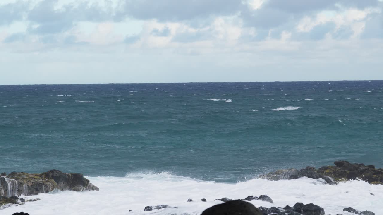 A dynamic coastal scene of turquoise ocean waves breaking with force against dark volcanic rocks, sending white spray into the air under a partly cloudy sky. Captures the raw power of nature