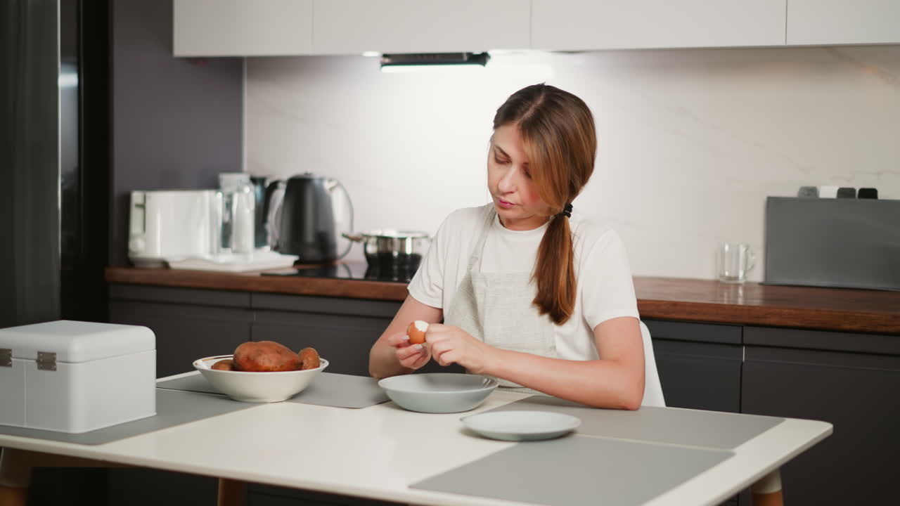 Woman wearing white shirt and apron seated at kitchen table peeling shell off boiled egg with head tilted to side, surrounded by plates, bowl of potatoes, and modern appliances in clean home kitchen
