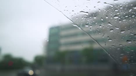 Water-running-down-the-windshield-of-the-car,-the-wipers-working-in-rain,-a-blurred-city-in-the-background