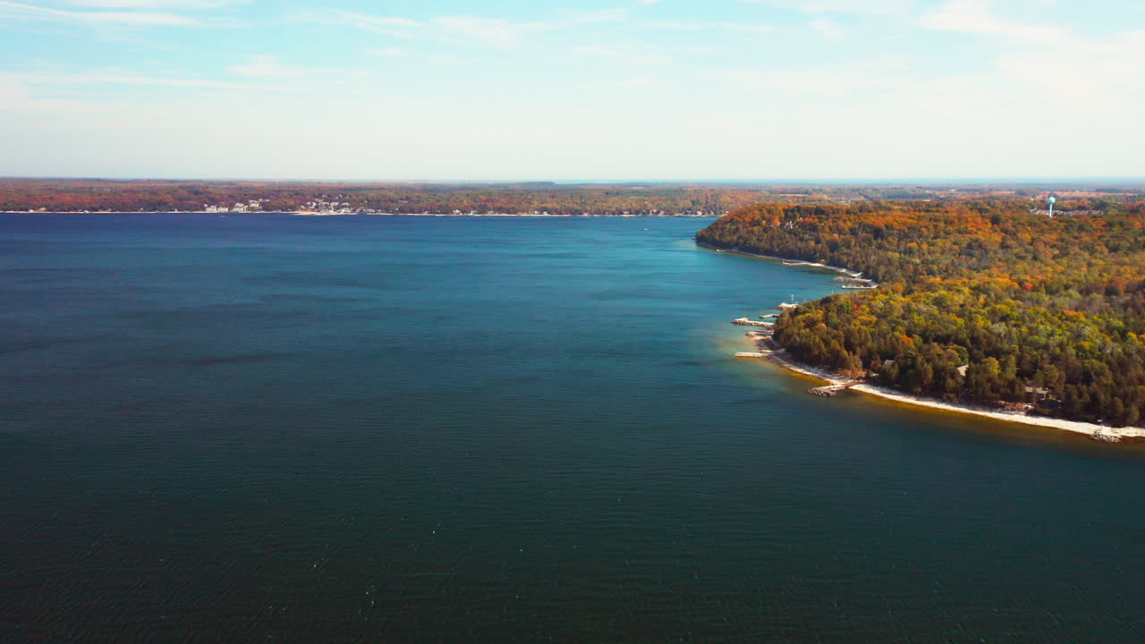 Vibrant autumn forests stretch along a sandy curve where the land meets deep blue water, forming a graceful coastline bathed in soft sunlight beneath open skies