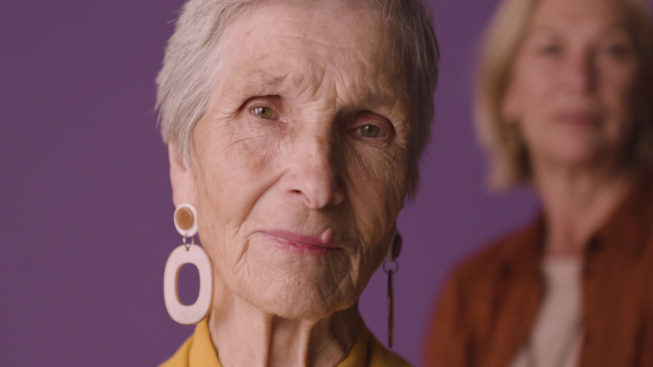 겨자색 셔츠와 재킷, 귀걸이를 하고 보라색 배경에 흐릿한 성숙한 여인과 함께 포즈를 취한 짧은 머리 금발의 노부인의 모습