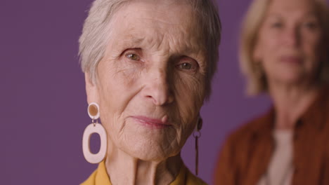 close up view of blonde senior woman with short hair wearing mustard colored shirt and jacket and earrings, posing with blurred mature woman on purple background