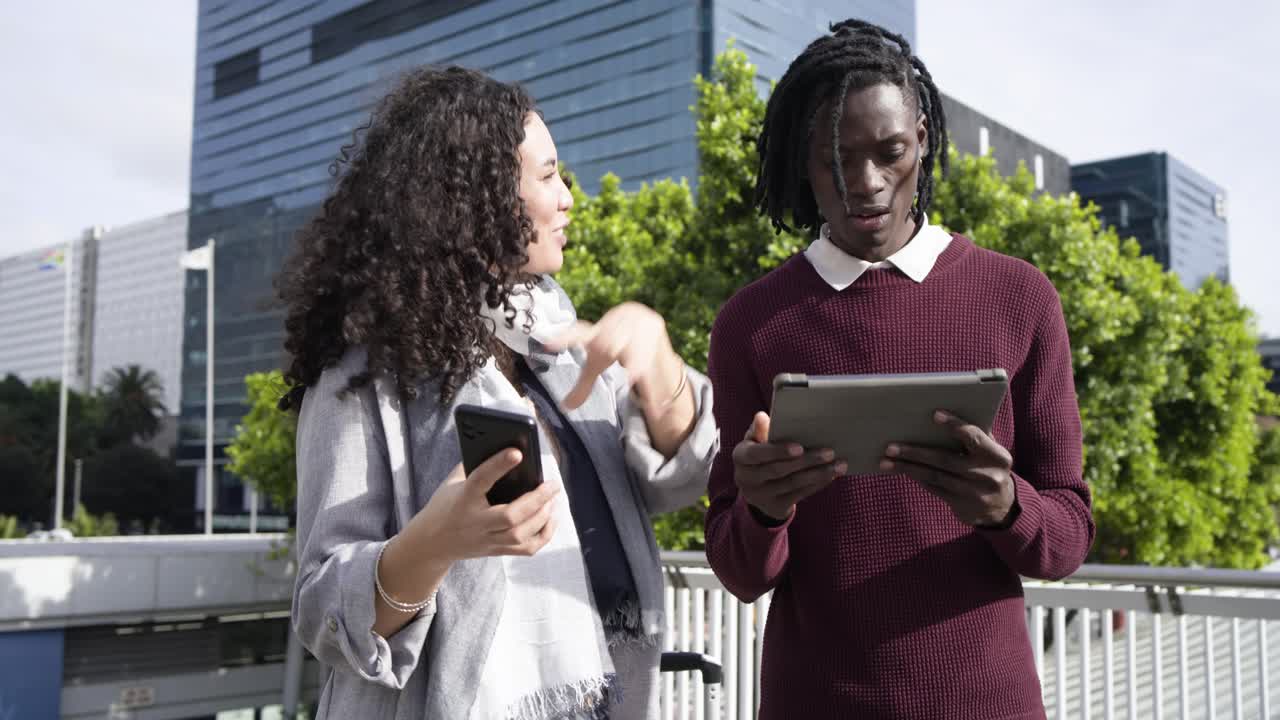 Diverse coworkers raising smartphone and tapping tablet outside blue-glass building and high-fiving