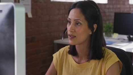 Mixed-race-businesswoman-wearing-headset-sitting-at-desk-using-computer-smiling