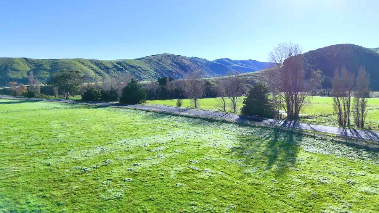 A car travels through vibrant green fields and rolling hills under clear blue skies in Akaroa, New Zealand