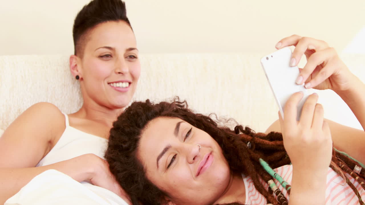 una pareja de lesbianas sonrientes usando un teléfono inteligente juntos en la cama