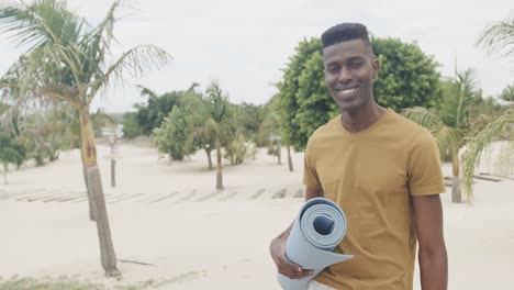 Portrait-of-happy-african-american-man-holding-rolled-up-yoga-mat-at-the-beach,-slow-motion