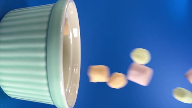 Marshmallows fall from ribbed ceramic cup against bright blue background creating playful food scene with motion and clean studio lighting