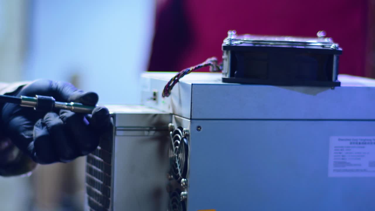 Close-up view of a technician performing maintenance on a cryptocurrency mining machine. The worker, wearing protective gloves, uses a tool to adjust or remove a cooling component from the miner