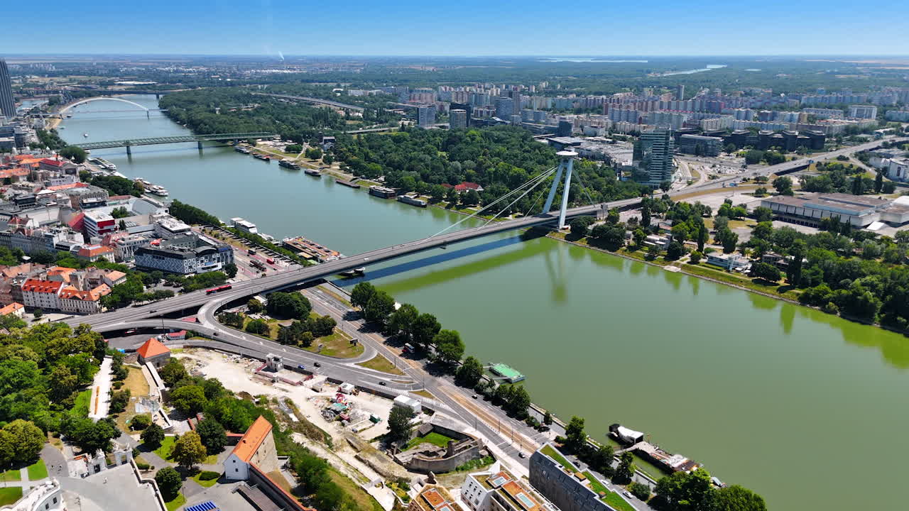 Footage over the waterfront of the Danube. Approaching the UFO Bridge over the river. Green cityscape of Bratislava at backdrop