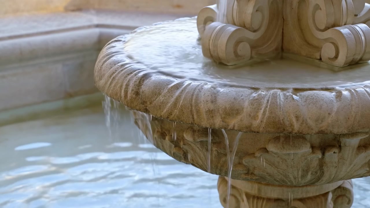 Primo piano di una vasca di fontana in pietra ornata con acqua che si riversa dolcemente in una pozza calma, ambientato su uno sfondo delicatamente sfocato e illuminato dalla calda luce solare naturale.
