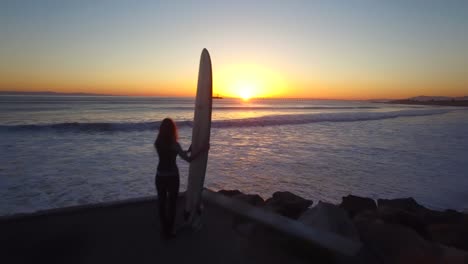 una antena sobre una mujer de pie con una tabla de surf mirando la puesta de sol 2