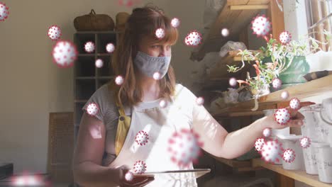 animation of virus cells over caucasian woman with face mask inspecting plants