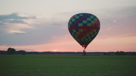 colorful checkerboard hot air balloon drifting low over vast green crop field at dusk under pastel sky glowing pink horizon in tranquil rural landscape