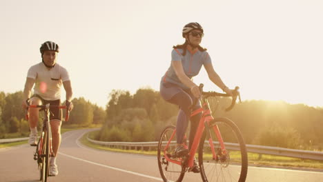 steadicam shot of two healthy mem and woman peddling fast with cycling road bicycle at sunset