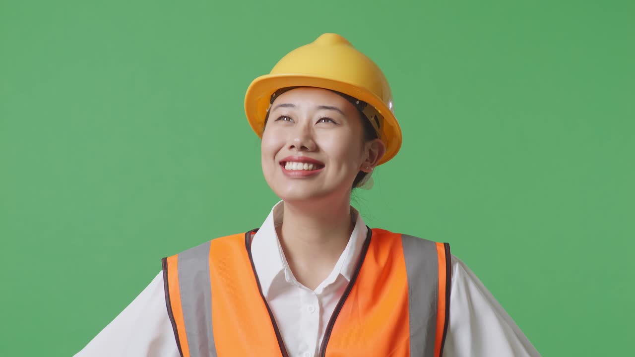 안전 헬을 착용 한 아시아 여성 엔지니어의 근접 사진은 녹색 스크린 배경 스튜디오에서 팔을 들고 서있는 동안 주위를 둘러보고 있습니다.