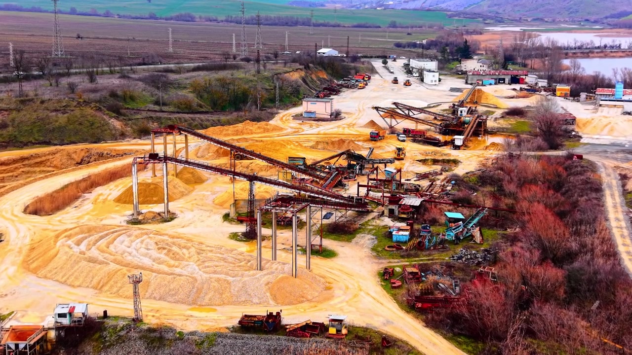 Overcast view of quarry complex and surrounding fields. Aerial perspective shows conveyors, stockpiles, power lines, and distant mountains.