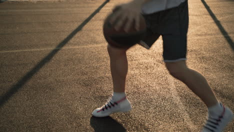 Primer-Plano-De-Un-Jugador-De-Baloncesto-Masculino-Rebotando-Y-Botando-La-Pelota-Entre-Sus-Piernas-En-Una-Cancha-Al-Aire-Libre-Al-Atardecer