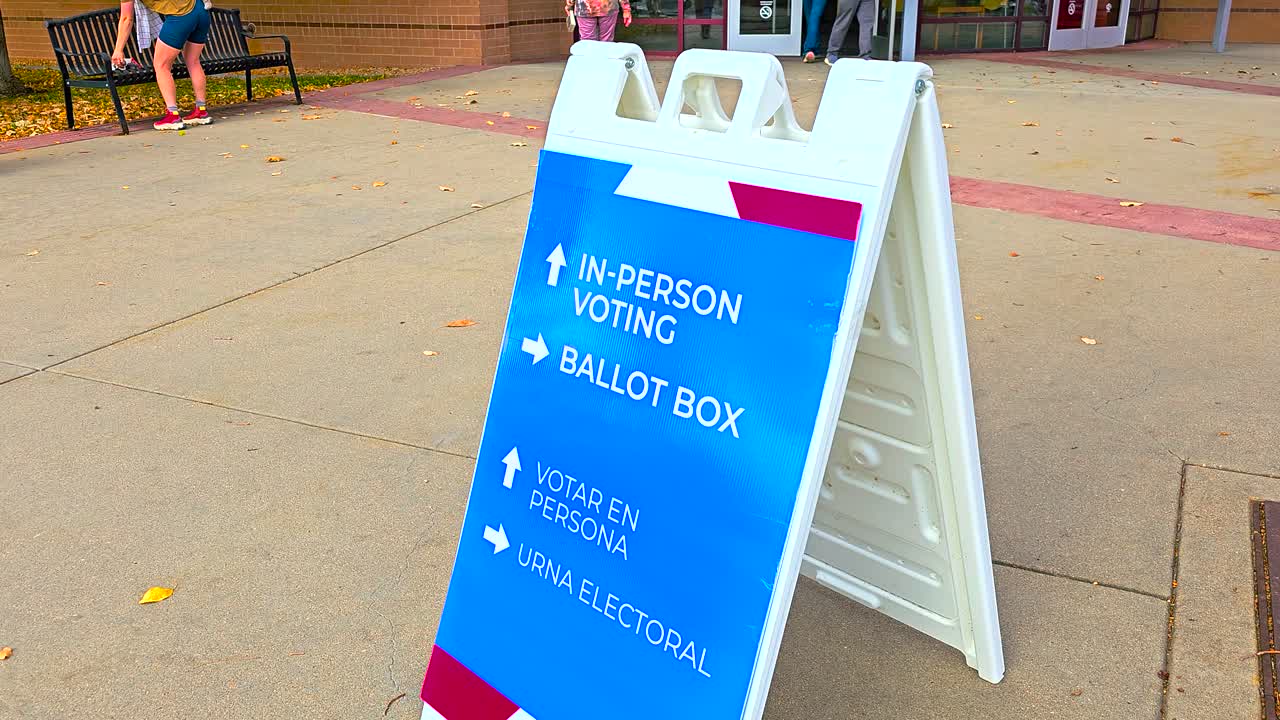 People walk in the background as a ballot drop-off sign with arrows displays Spanish words Votar en Persona and Urna Electoral, symbolizing civic duty, inclusivity, democracy, and secure participation