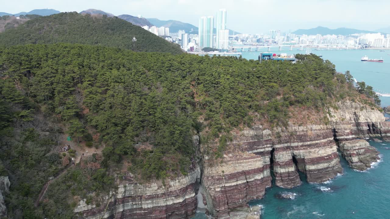 Drone aerial view in South Korea countryside flying over the clear blue sea of Busan next to a green mountain, ships and city in the background sunny day