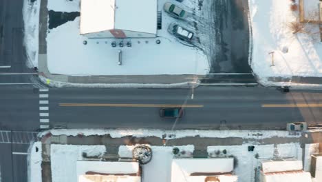 top down aerial tracking shot of black car driving through snow covered town in winter during magic hour light