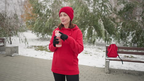 middle-aged woman in red hoodie and beanie opens water bottle outdoors, holding it while standing in snow-dusted park with red bag on bench, surrounded by trees and serene winter atmosphere