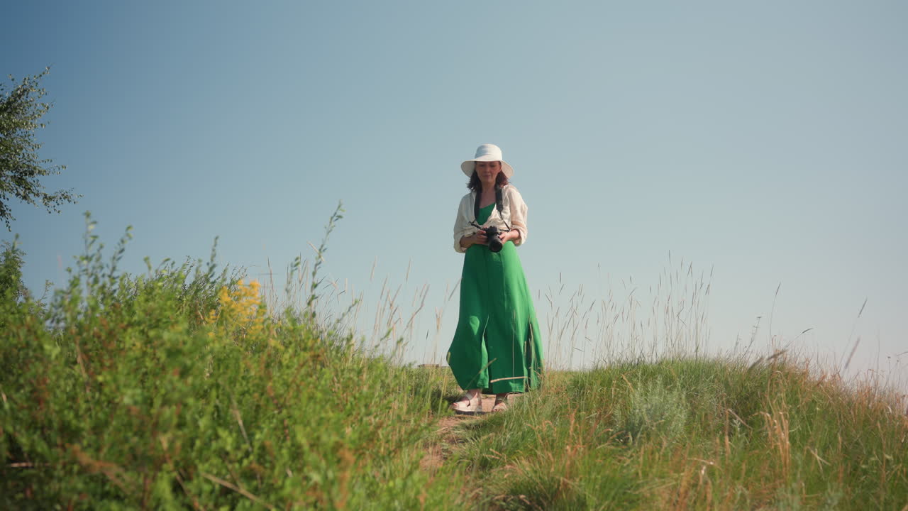 Una turista che indossa un cappello da sole e un vestito verde tiene in mano una macchina fotografica mentre scende un sentiero erboso in cima a una collina in una giornata di sole, circondata da erba alta, fiori selvatici e un cielo luminoso.
